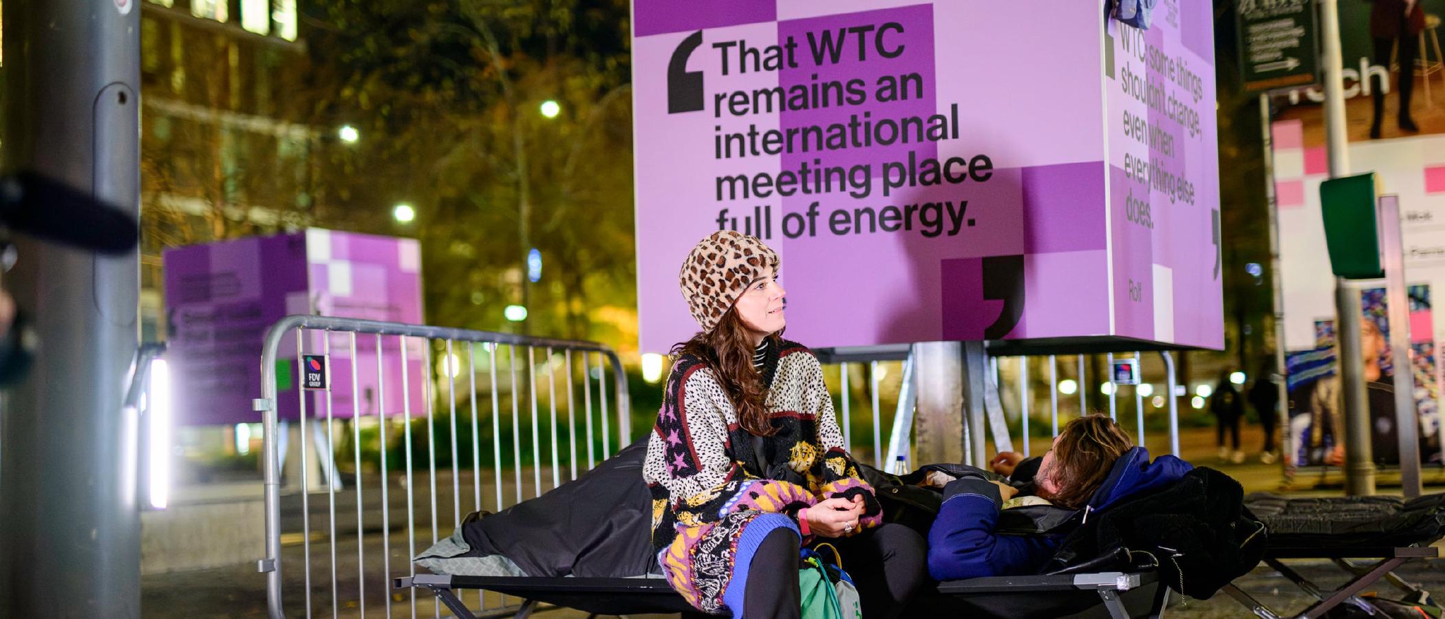 Bezoeker in gesprek naast een campingbed en een WTC-billboard tijdens de Sleep Out voor Sheltersuit