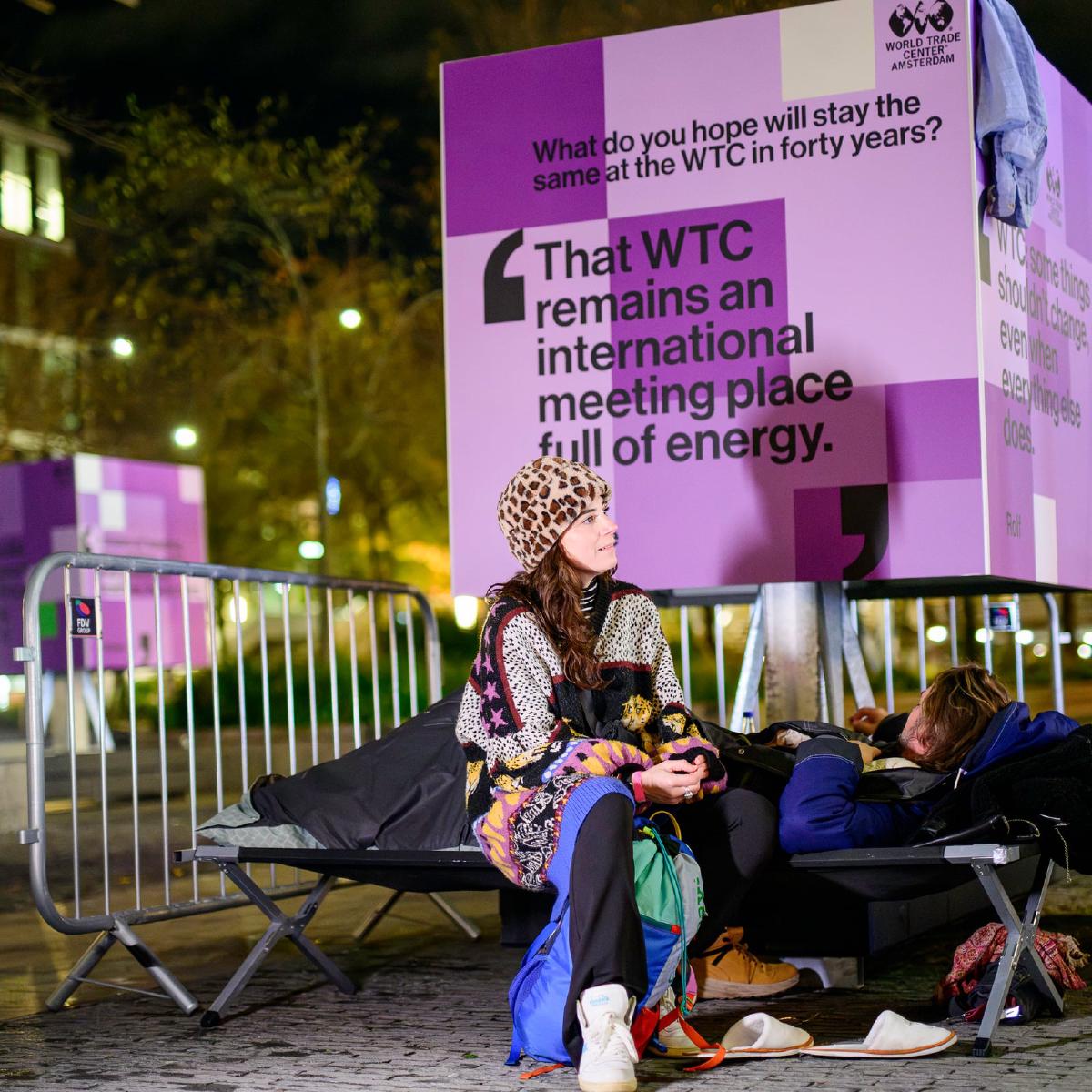 Bezoeker in gesprek naast een campingbed en een WTC-billboard tijdens de Sleep Out voor Sheltersuit
